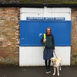 Angela standing with Flynn her guide dog, in front of a huge blue noticeboard
