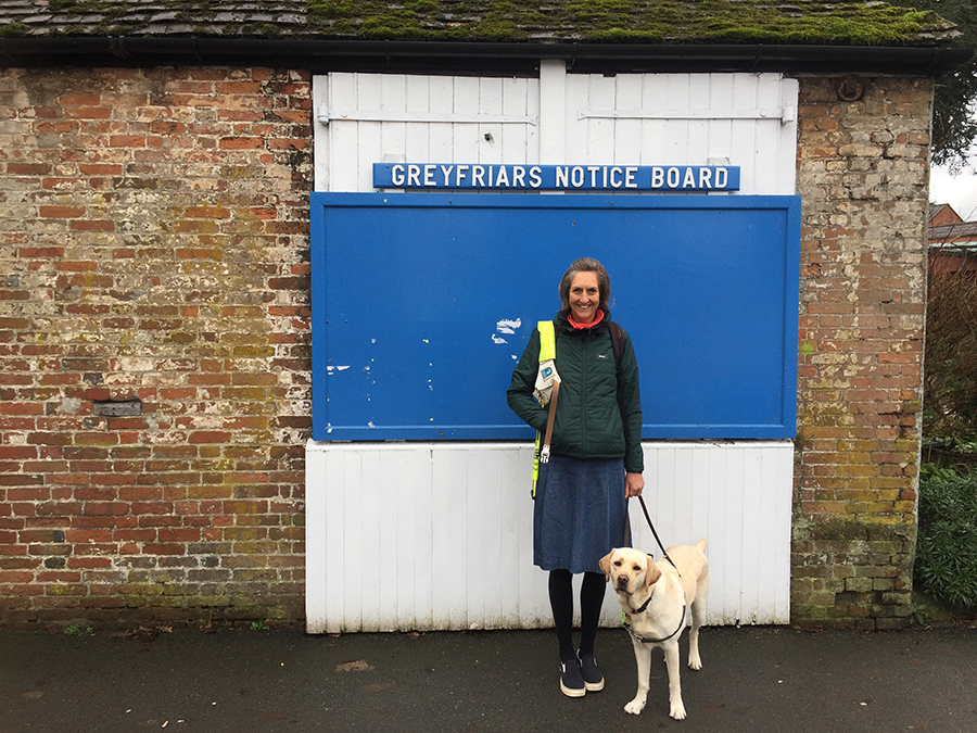 Angela standing with Flynn her guide dog, in front of a huge blue noticeboard