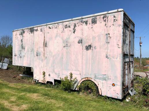 Disused, faded pink removal van body with no cab. Brambles growing out of where the wheels once were