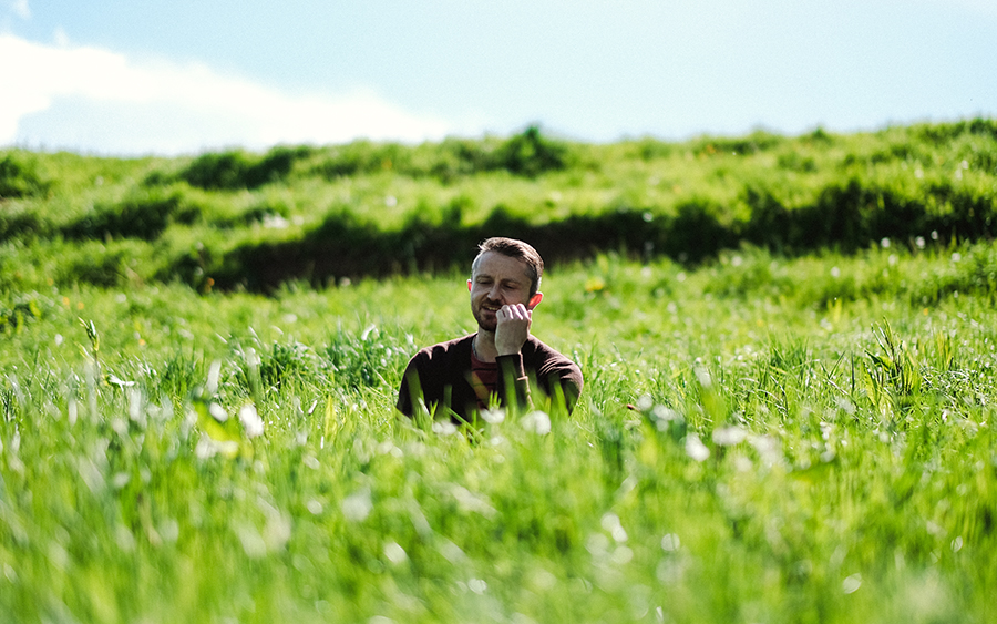 Phil Owen sitting in long grass, singing his hand is on his chin