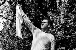 Xavier wearing a pale jumper, standing with a serious expression on his face with his right hand in the air holding a white flag, tress in the background