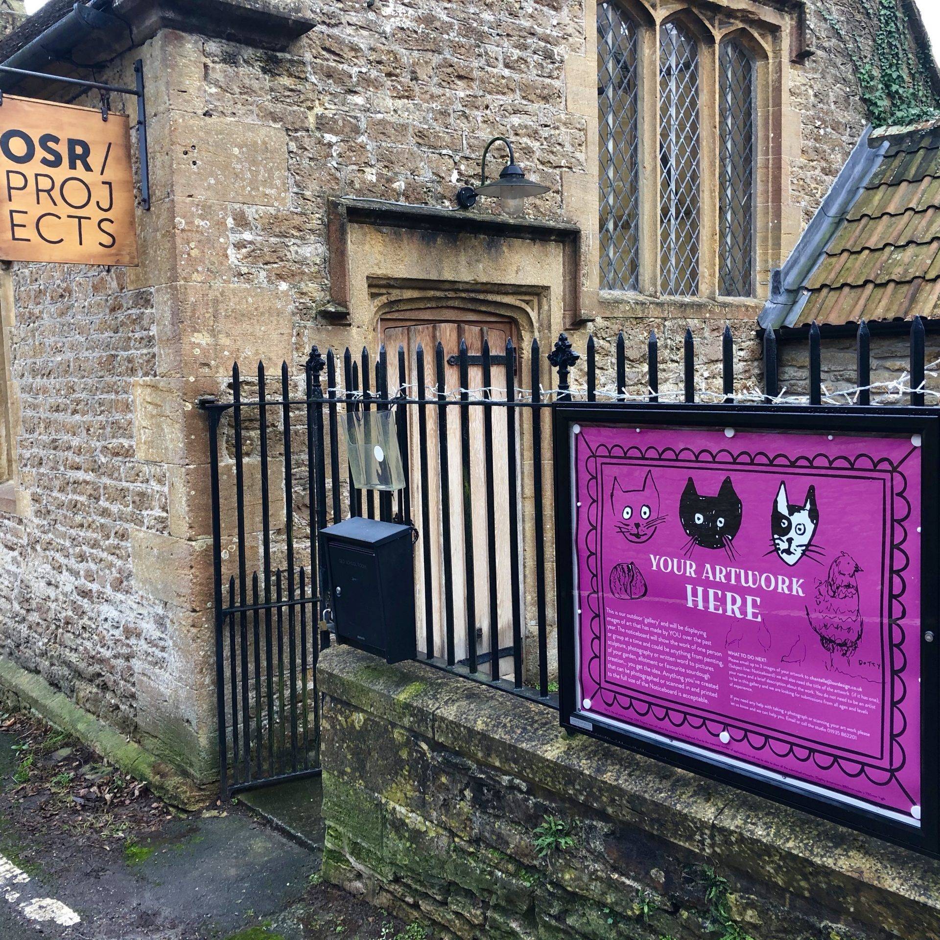 Noticeboard attached to the railings of OSR Projects. Inside the noticeboard is a pink lare poster with sketches and wording your artwork here.