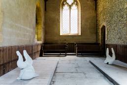 two grey concrete sculptures, child like bodies by tania kovats in a chapel with light streaming through the window