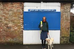 angela wearing a green coat and blue skirt with flynn by her side, standing in front of a white wooden garage door with a blue noticeboard