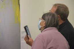 two people wearing face masks holding their phone near their faces whilst looking closely at a painting