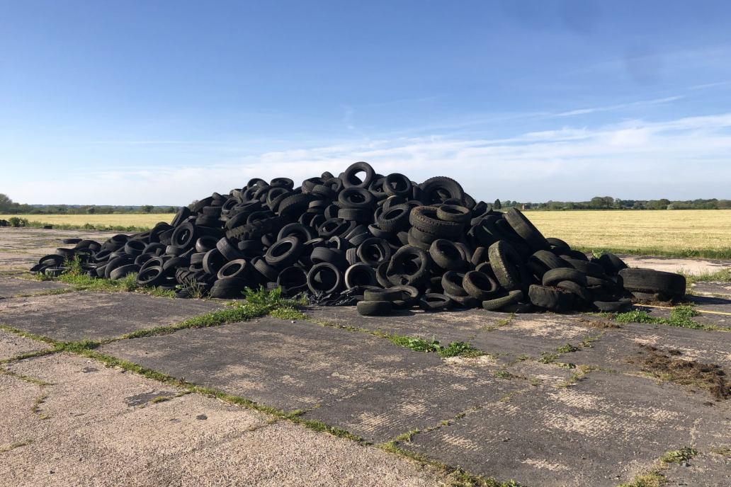a huge pile of old tyres on a concrete surface on the edge of a field with blue sky