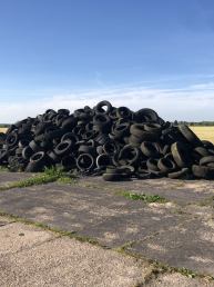 a huge pile of old tyres on a concrete surface on the edge of a field with blue sky