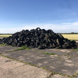 a huge pile of old tyres on a concrete surface on the edge of a field with blue sky