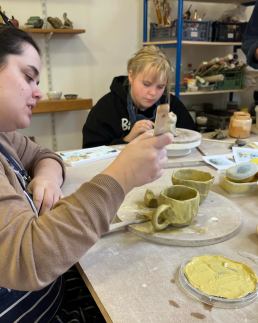 two people making with clay, one decorating a pinch pot cup with yellow slip and the second person decorating a clay cat