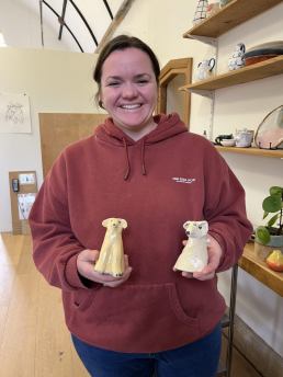 a smiling person, wearing a burgandy hoody, holding two ceramic yellow dogs