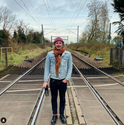 scoty standing on a train track wearing a denimin jacket, black jeans, red scarf and beanie