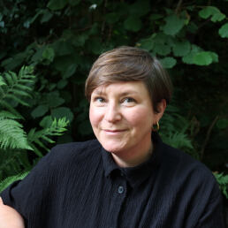 susie looking directly at the camera, she has short brown hair and is wearing a black collared shirt. There are green ferns behind her.