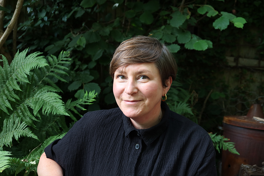 susie looking directly at the camera, she has short brown hair and is wearing a black collared shirt. There are green ferns behind her.